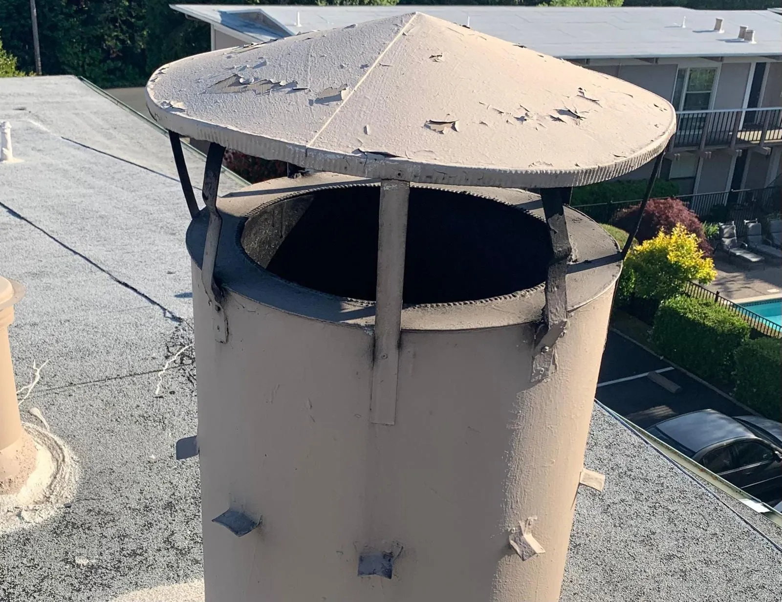 A round metal chimney cap with chipped beige paint over an open flue on a flat roof, with an apartment building and a swimming pool behind it